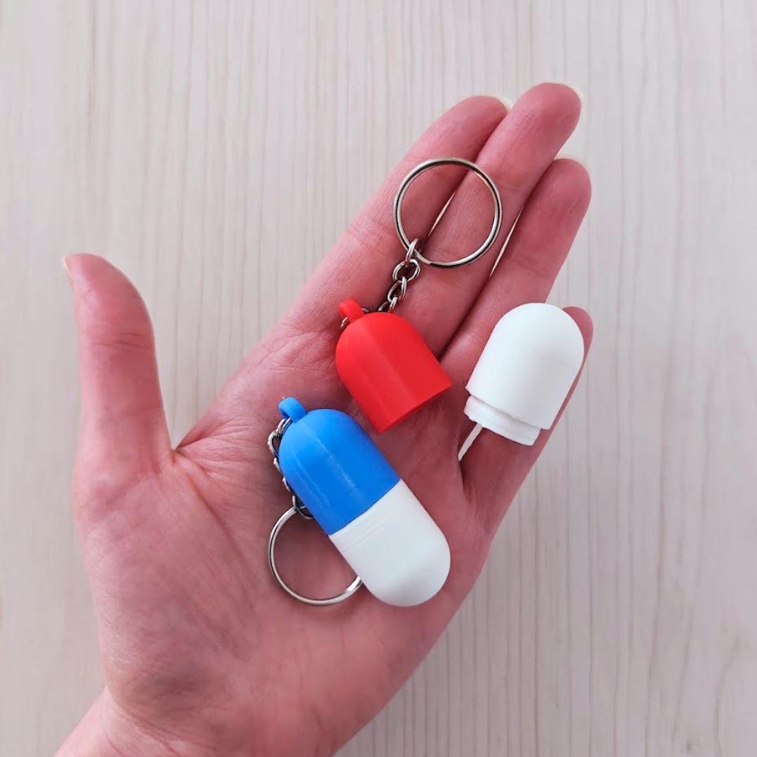 Hand holding three miniature capsule keychain containers in red, blue and white, showing their compact size.