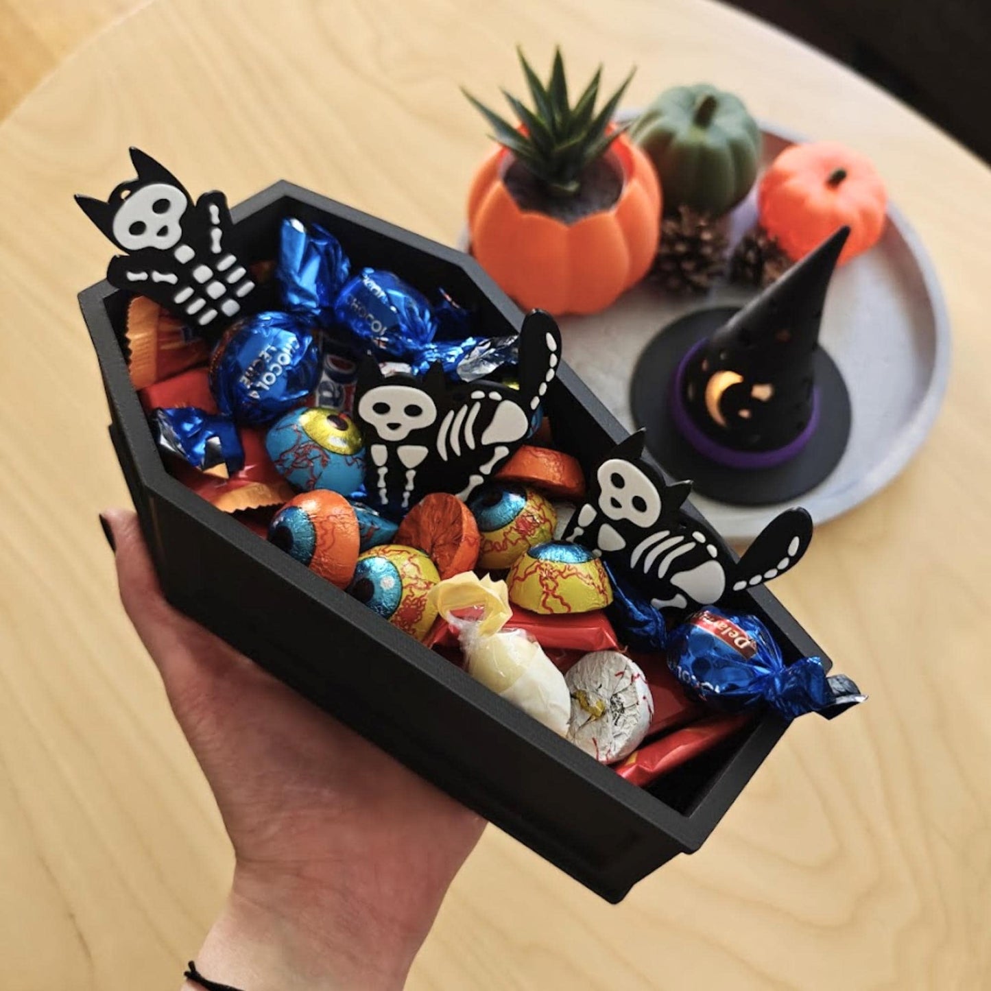 Coffin candy bowl held in hand, filled with assorted Halloween candies and surrounded by pumpkins and decor.