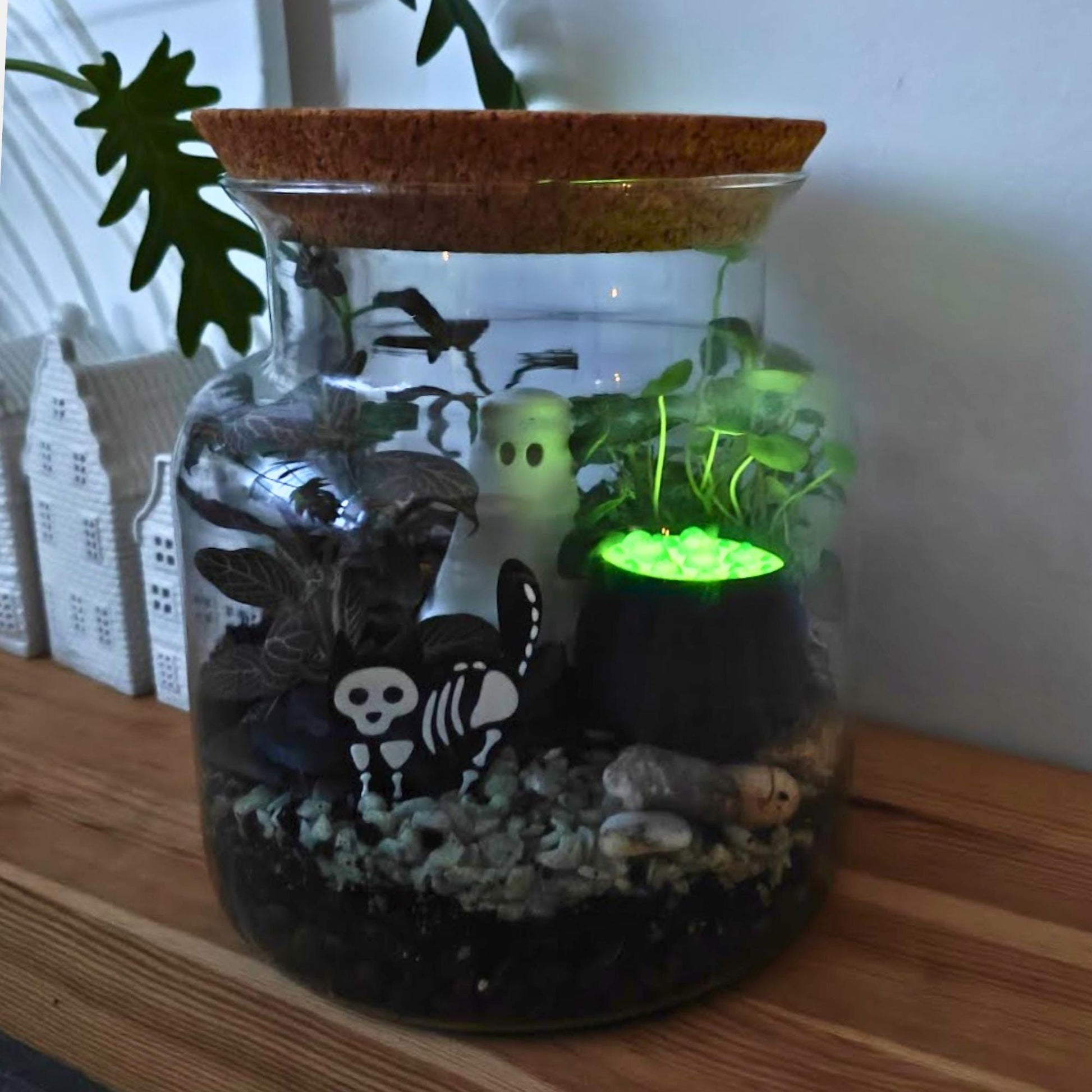 Halloween terrarium display featuring the glowing green cauldron among plants, skulls, and small skeleton decorations.