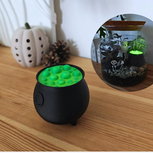 Black 3D-printed cauldron tealight holder with glowing green bubble top, displayed on a wooden shelf beside Halloween pumpkins and mini houses