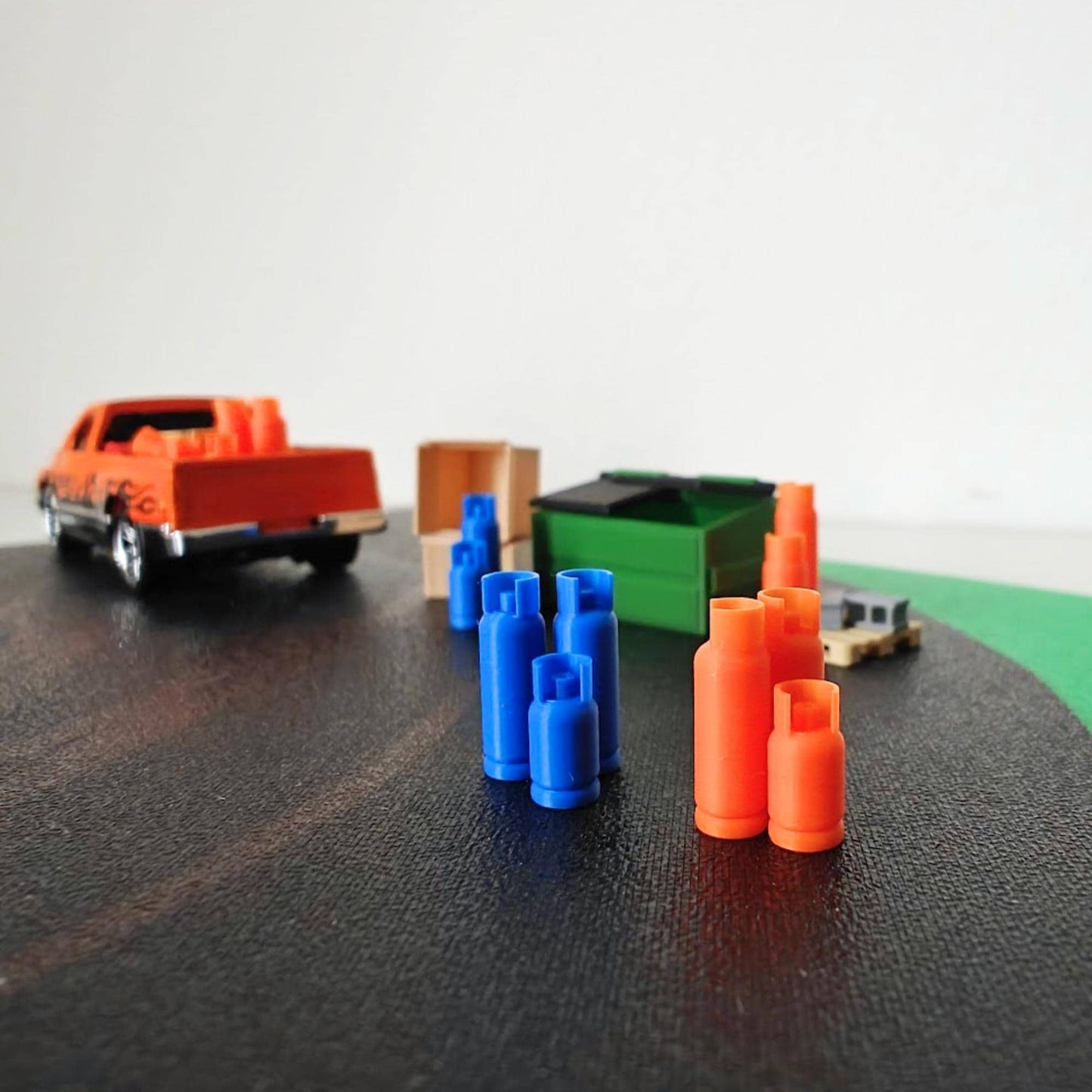 Miniature 1:64 scale scene featuring blue and orange 3D-printed gas bottles arranged near a green dumpster, wooden crate, and pickup truck on a black diorama surface.