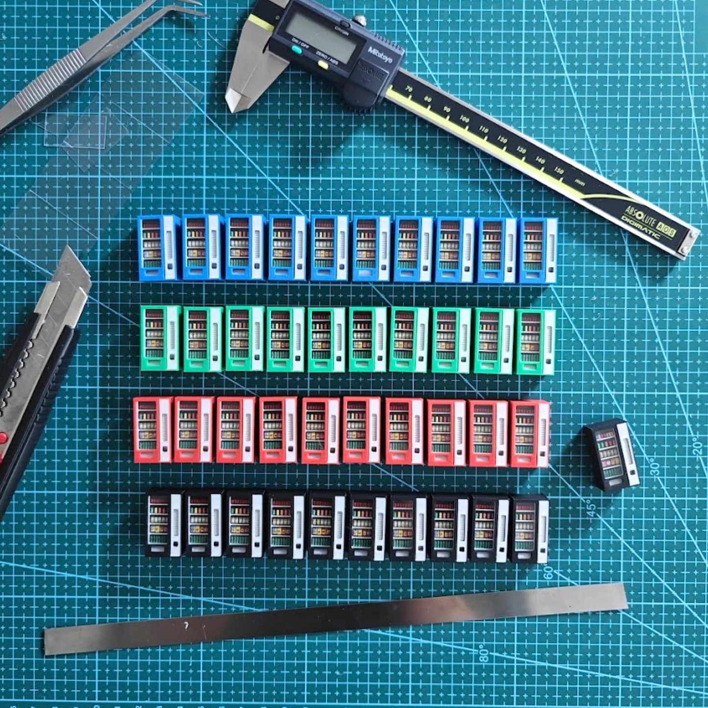 Rows of 3D-printed miniature vending machines in red, blue, green, and black lined up on a cutting mat, showing painting and detailing process.