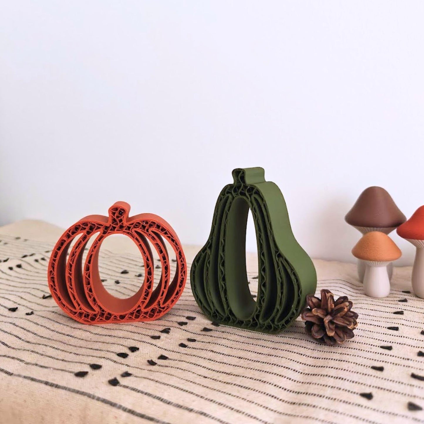 3D-printed pumpkin and gourd decorations in terracotta orange and dark green displayed on a fall-themed table with pinecones and acorns.