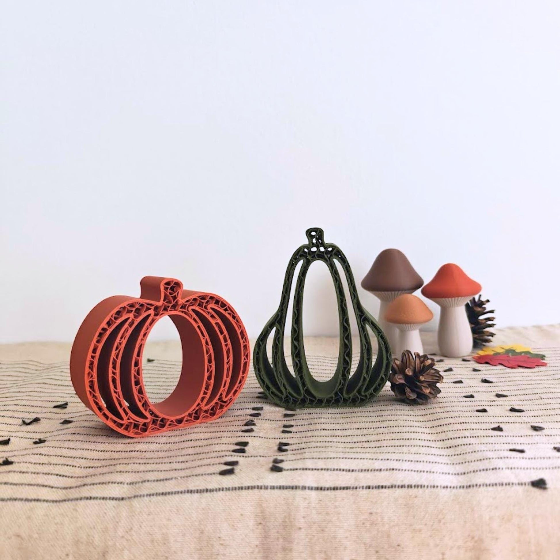 Close-up of orange and green 3D-printed pumpkins and gourds with small decorative leaves on a beige woven cloth.
