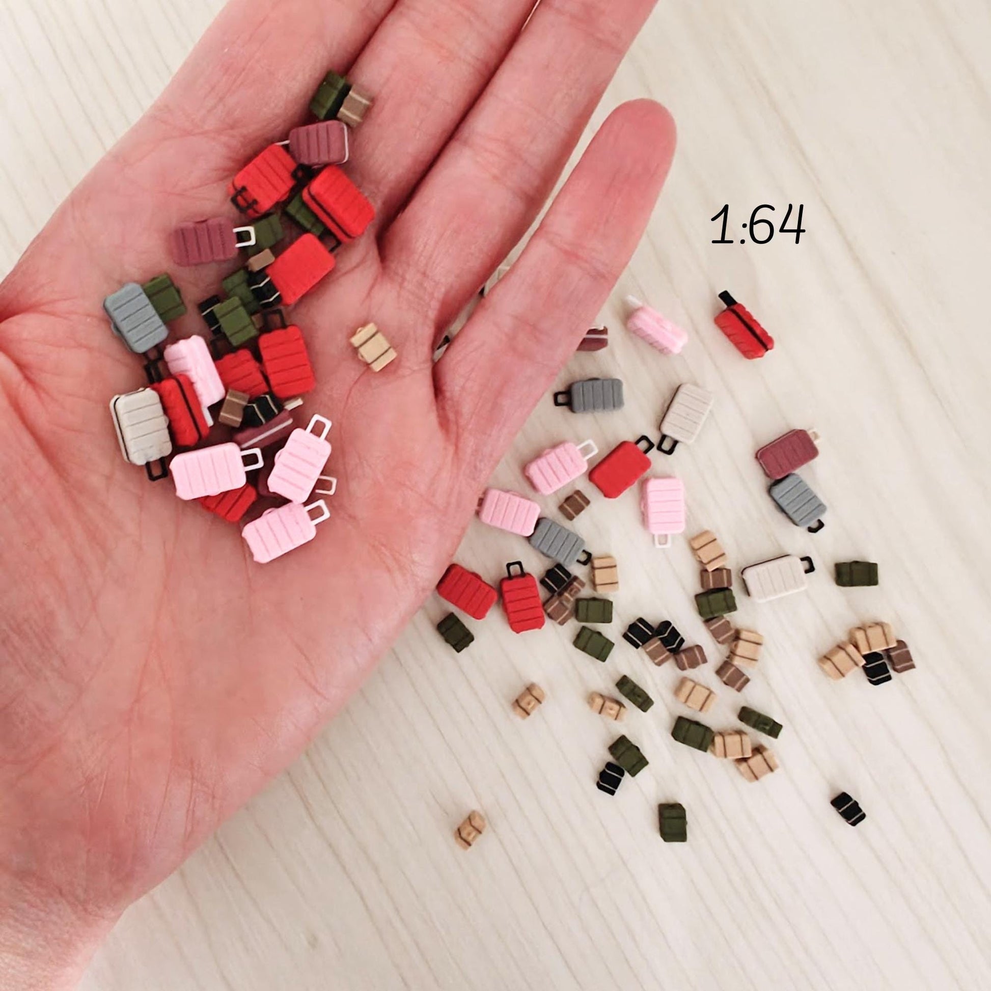 Miniature 3D-printed suitcases in scale 1:64, held in a person’s hand for size reference. Includes various colors such as red, pink, green, and brown.