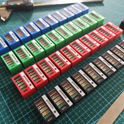 Rows of 3D-printed miniature vending machines in red, blue, green, and black lined up on a cutting mat, showing painting and detailing process.