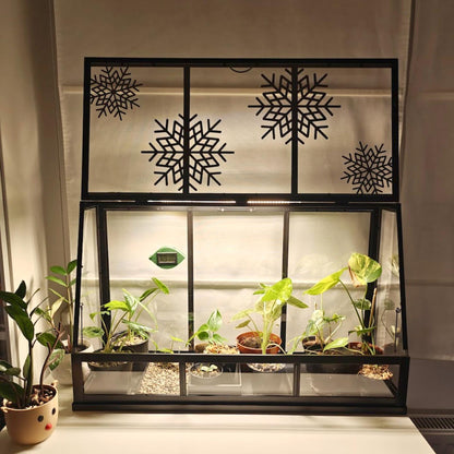Open black Äkerbar greenhouse with snowflake decorations, placed on white dresser near potted plant.