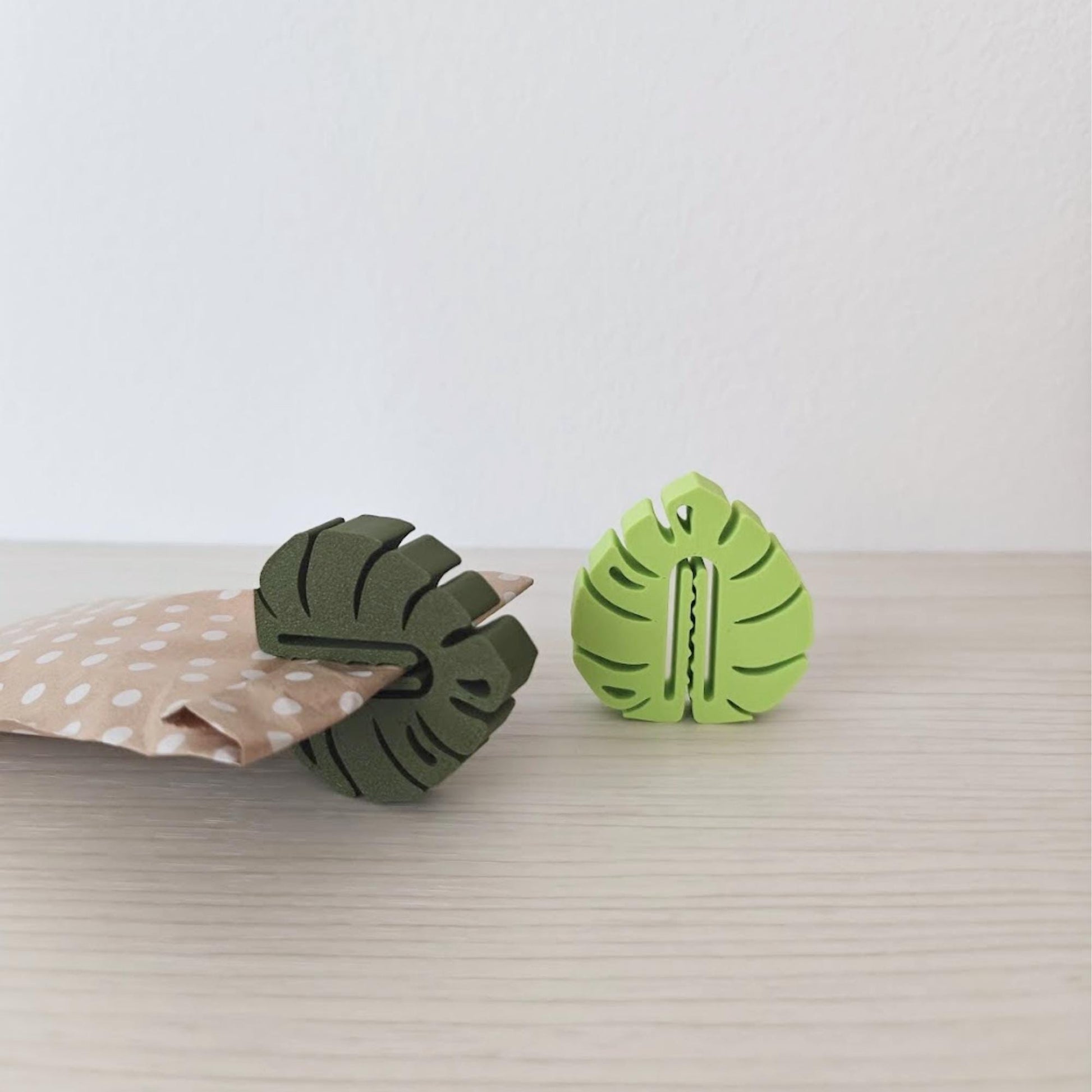 Green monstera leaf clips displayed with a small kraft paper pouch on a light tabletop.