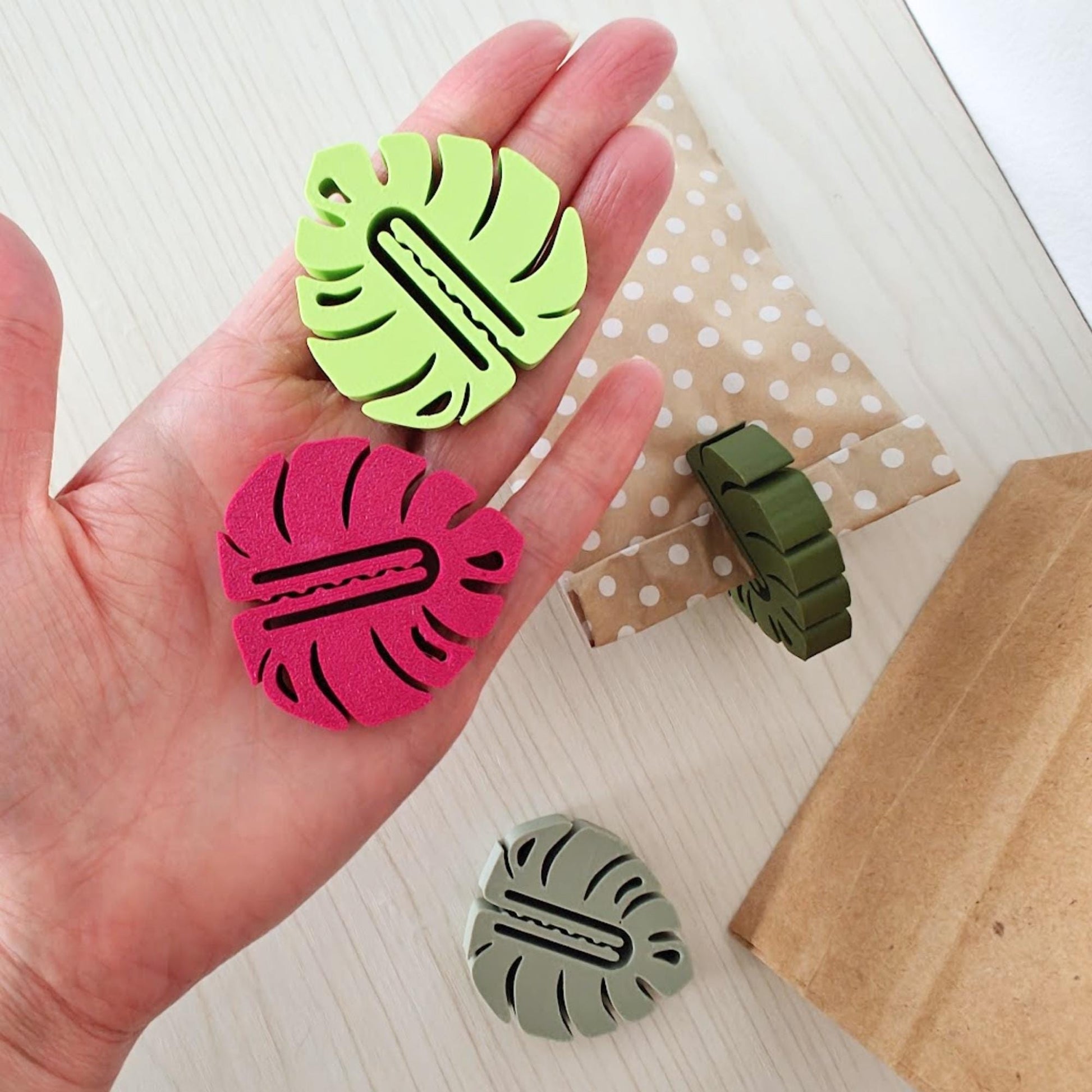 Five colorful monstera leaf clips held in hand with kraft paper packaging in the background.