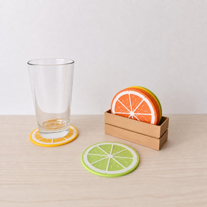 Citrus coaster set with a glass placed on a lemon coaster, lime coaster on the table, and orange coasters stored in a wooden crate.