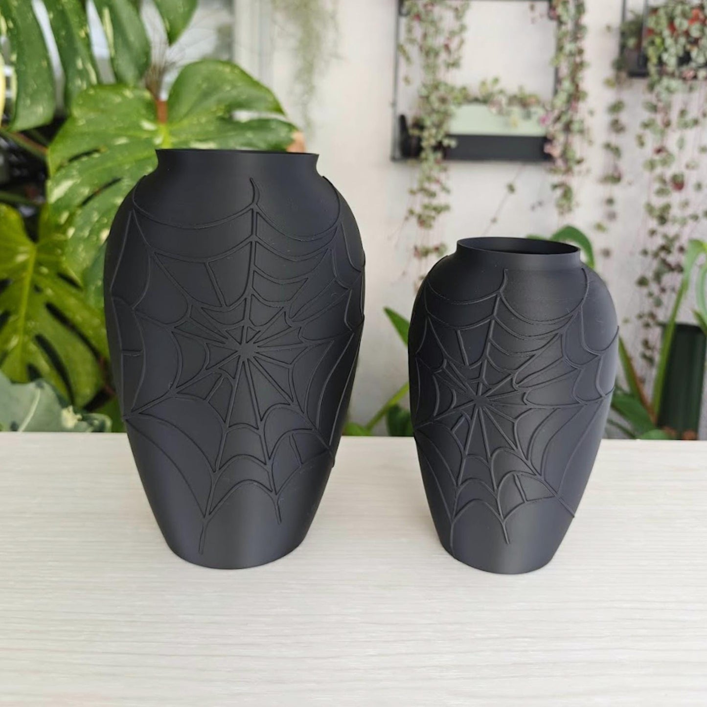 Black spider web vases in two sizes displayed on a light shelf with green plants in the background.