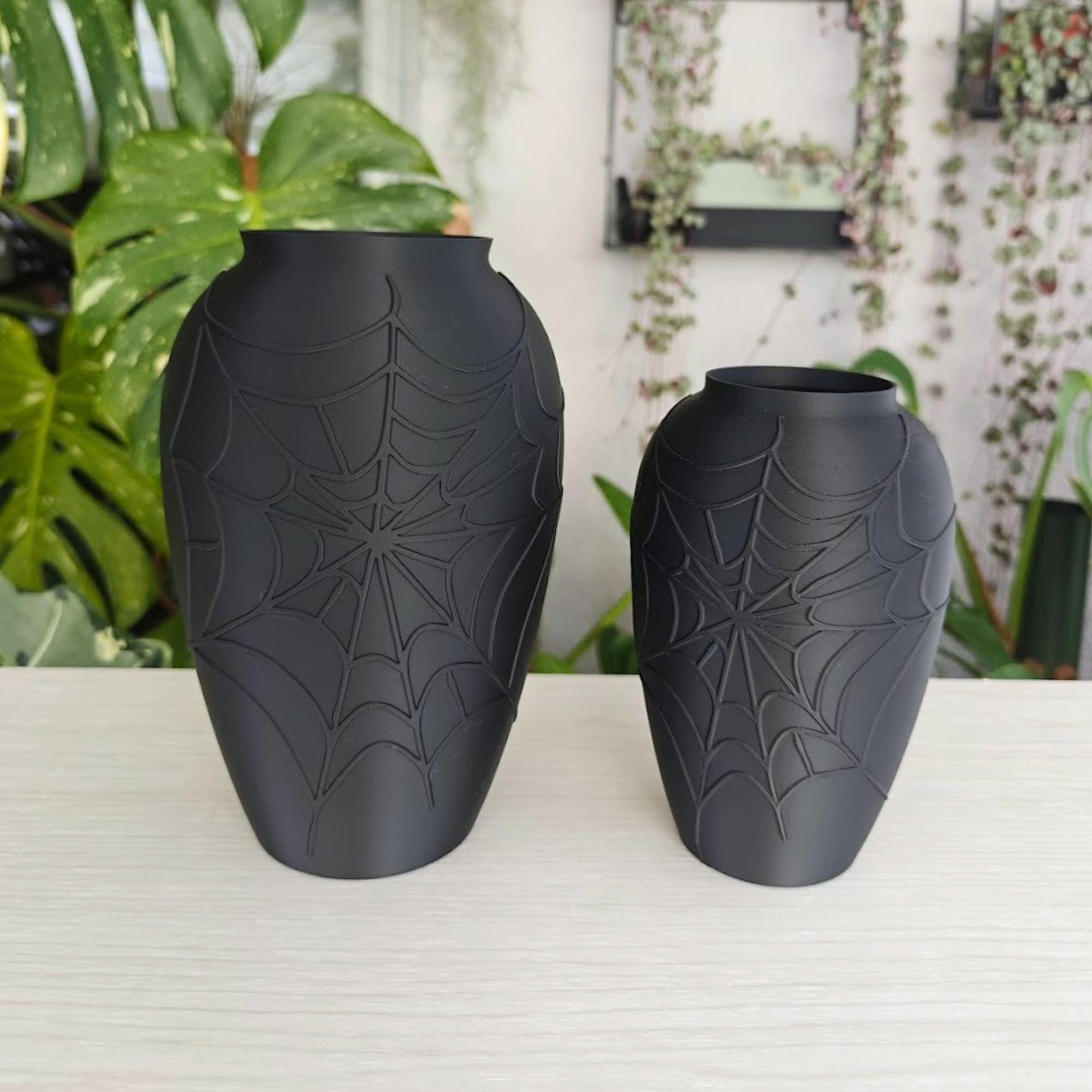 Black spider web vases in two sizes displayed on a light shelf with green plants in the background.