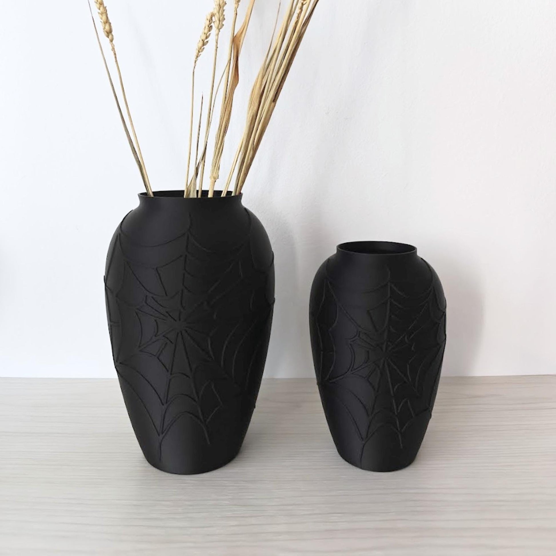 Two black spider web vases displayed with dried flowers on a light wooden shelf.