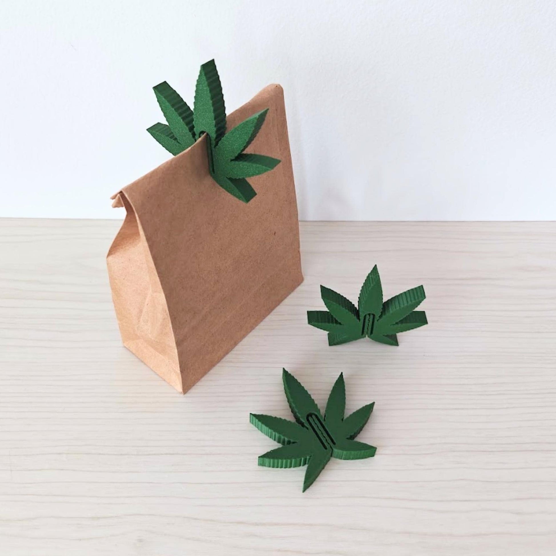 Three marijuana leaf-shaped food clips shown beside a small kraft paper bag against a neutral background.