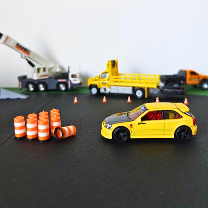 Miniature traffic barrels placed around a yellow model car in a roadwork diorama with cranes and construction vehicles.