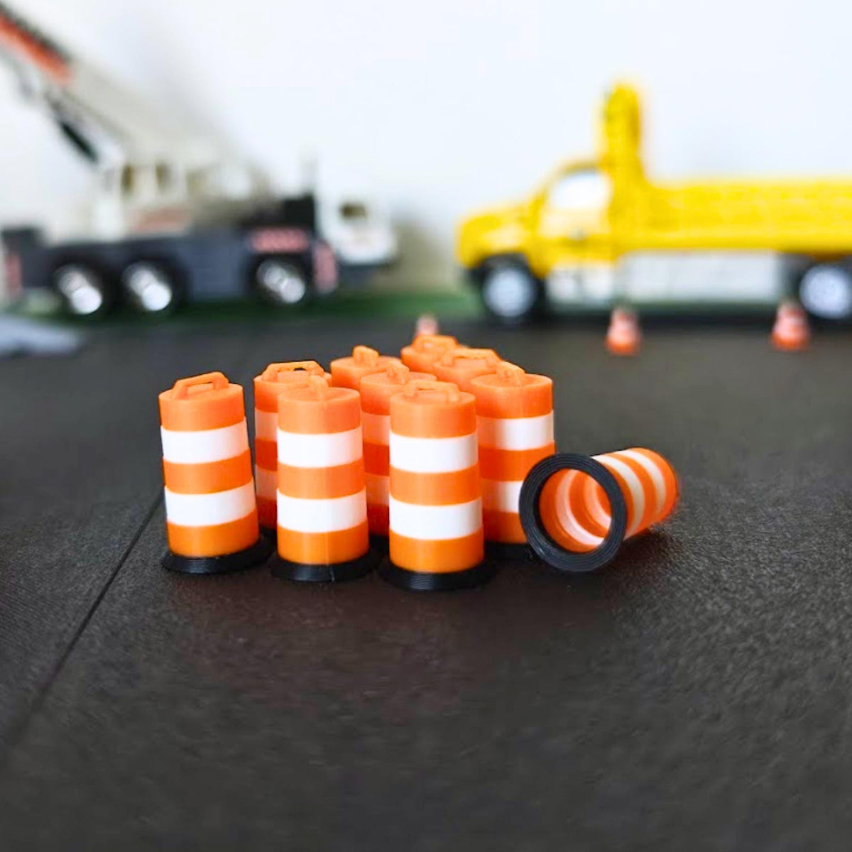 Set of miniature orange traffic barrels with white bands arranged in front of a scale crane and work truck.