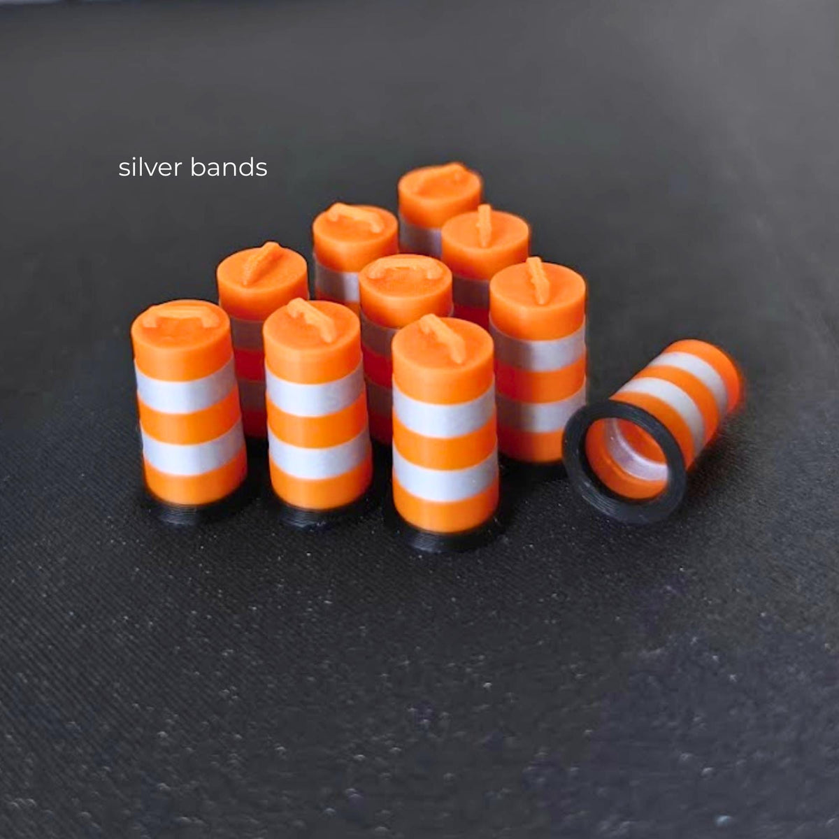 Ten miniature orange traffic barrels with metallic silver bands and black bases on a dark background.