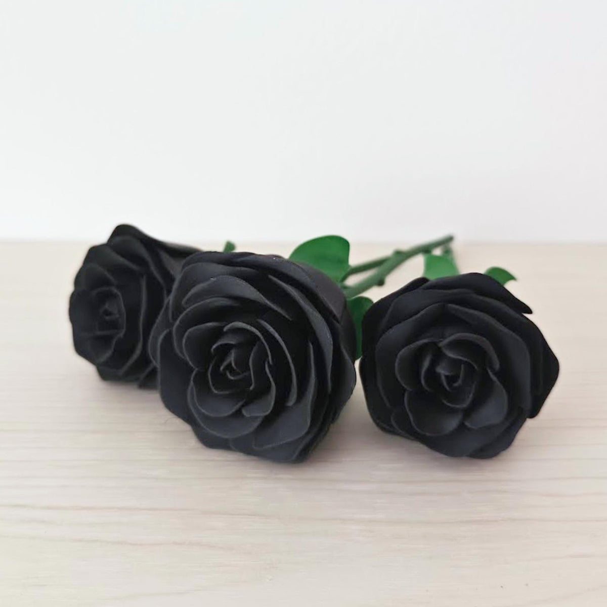 Three matte black rose heads arranged on a light wooden surface with green stems visible behind them
