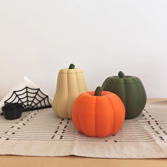 Three lidded pumpkin storage containers in different shapes and colors displayed with black spiderweb Halloween decorations.